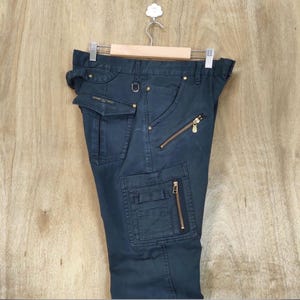 Pu&ograve; includere: Pantaloni cargo blu scuro con multiple tasche e cerniere dorate. I pantaloni sono appesi a una gruccia di legno su uno sfondo marrone chiaro. I pantaloni hanno un design utilitario con un anello a D e una piccola tasca.