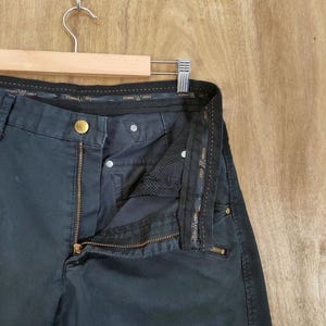 Pu&ograve; includere: Pantaloni da golf grigio scuro con cerniera in ottone parzialmente aperta e bottone dorato. I pantaloni sono appesi a una gruccia di legno su uno sfondo di legno marrone chiaro. Il marchio "EDWIN GOLF" &egrave; visibile.