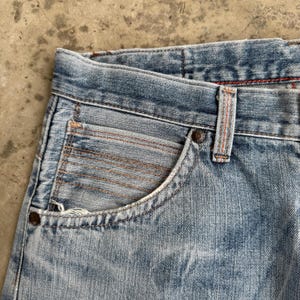May include: Close-up of light blue denim jeans. The jeans have a classic five-pocket design with a small pocket on the front. The stitching is a contrasting orange color, and the jeans appear to be worn and faded.