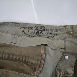 May include: Close-up of khaki-colored pants with a Route 66 graphic and the text "Get Your Kicks, Main St, America". The pants feature metal button and rivet details.