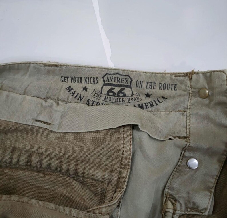 May include: Close-up of khaki-colored pants with a Route 66 graphic and the text "Get Your Kicks, Main St, America". The pants feature metal button and rivet details.