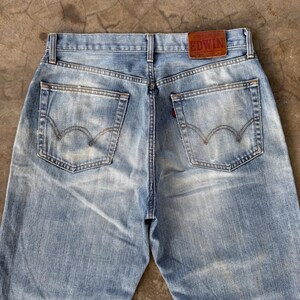 May include: Light blue denim jeans with a faded wash and classic five-pocket design. Features a brown leather patch with the brand name "EDWIN" on the back waistband. The jeans have a straight leg cut and are pre-worn.