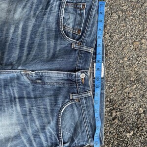 May include: Close-up of blue denim jeans with a faded wash. A blue measuring tape shows the jeans are approximately 21 inches long. The jeans have classic five-pocket styling and a button closure.