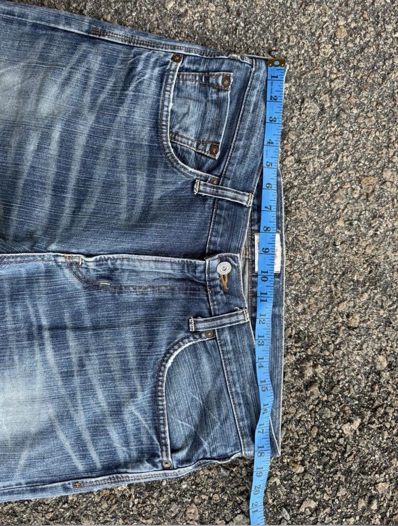 May include: Close-up of blue denim jeans with a faded wash. A blue measuring tape shows the jeans are approximately 21 inches long. The jeans have classic five-pocket styling and a button closure.