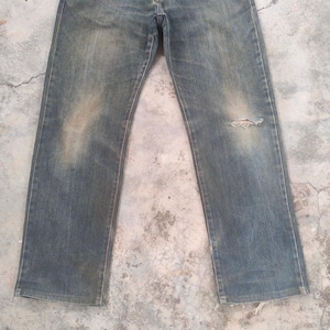 May include: Faded blue denim jeans with a distressed appearance. The jeans have a straight leg cut and show signs of wear, including a small tear on the right knee area. The denim has a worn, vintage look.