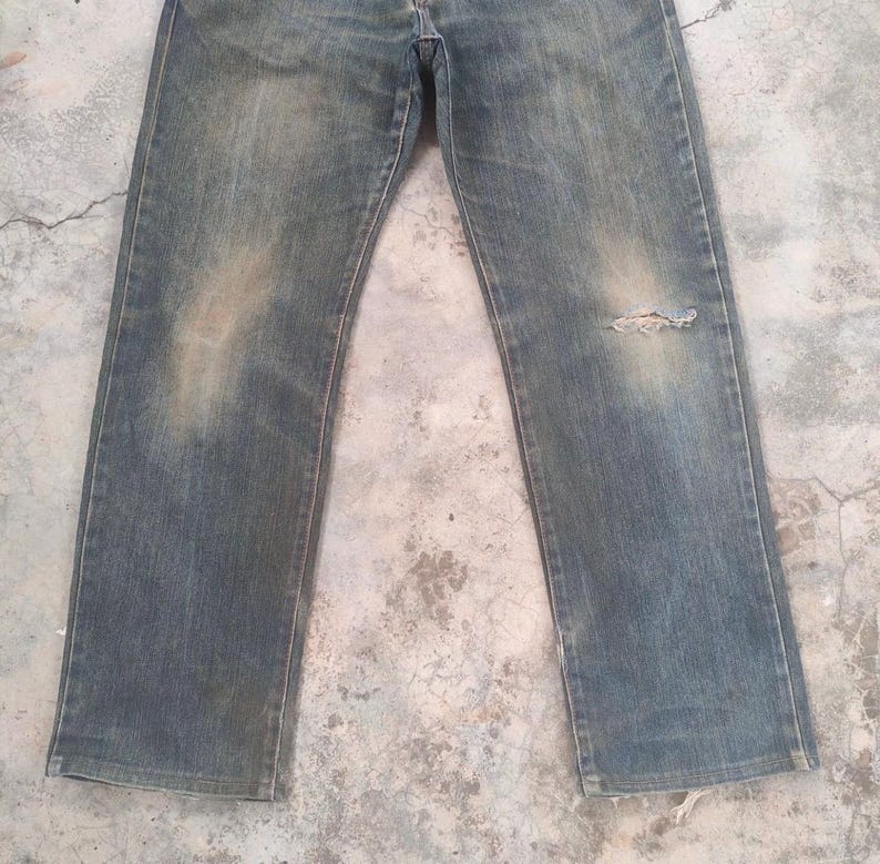 May include: Faded blue denim jeans with a distressed appearance. The jeans have a straight leg cut and show signs of wear, including a small tear on the right knee area. The denim has a worn, vintage look.