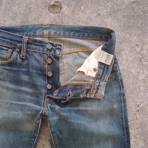 May include: Faded blue denim jeans with a button fly. The jeans are unbuttoned, revealing the interior and a white tag. The denim has a worn appearance with visible fading and distressing.
