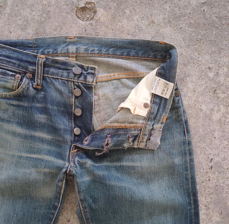 May include: Faded blue denim jeans with a button fly. The jeans are unbuttoned, revealing the interior and a white tag. The denim has a worn appearance with visible fading and distressing.