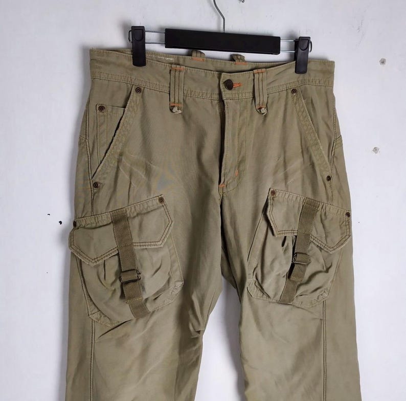 May include: Khaki-colored cargo pants with multiple pockets and straps. The pants have a button and zipper closure, belt loops, and orange stitching details. The pants are hanging on a black hanger against a white background.