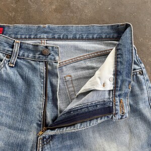 May include: Close-up of light blue denim jeans with a partially unzipped zipper. The jeans have a button closure and belt loops. The fabric shows signs of wear and fading, typical of denim.