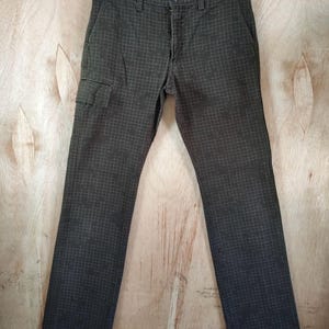 May include: Dark grey trousers with a grid pattern, featuring a cargo pocket on the left leg. The trousers are buttoned and zipped, hanging on a wooden hanger against a light wood background. The fabric appears to be hard-wearing.