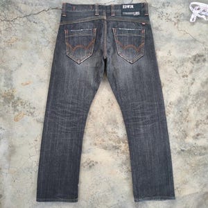 Pu&ograve; includere: Jeans in denim grigio scuro con dettagli di cuciture arancioni. I jeans hanno un classico design a cinque tasche e il nome del marchio "EDWIN" sul retro. I jeans sono appoggiati su una superficie di cemento.