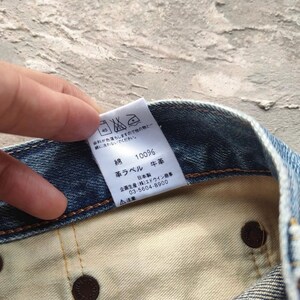 May include: Close-up of a denim garment with a white tag. The tag displays Japanese text, including "100% cotton" and washing instructions. The garment's interior is visible, showing brown rivets and a light-colored fabric.