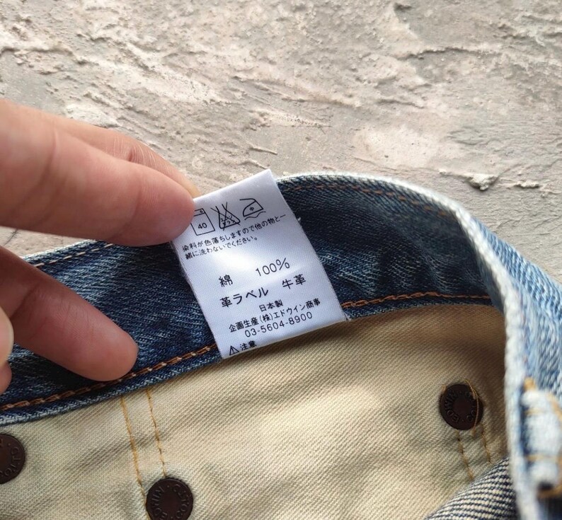 May include: Close-up of a denim garment with a white tag. The tag displays Japanese text, including "100% cotton" and washing instructions. The garment's interior is visible, showing brown rivets and a light-colored fabric.