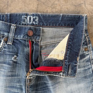 May include: Close-up of blue denim jeans with the number "503" printed on the waistband. The jeans have a red zipper and a button closure. The fly is partially unzipped, revealing the interior lining.