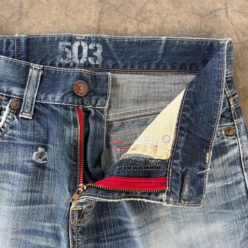May include: Close-up of blue denim jeans with the number "503" printed on the waistband. The jeans have a red zipper and a button closure. The fly is partially unzipped, revealing the interior lining.