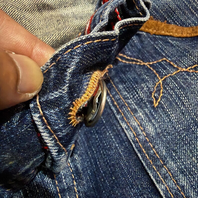 Op de afbeelding: Close-up van een denim kledingstuk, met een zilveren knoop en gedetailleerde stiksels. De blauwe stof heeft een versleten uitstraling, met oranje en rode draadaccenten. De knoop is vastgezet met goudgele draad.