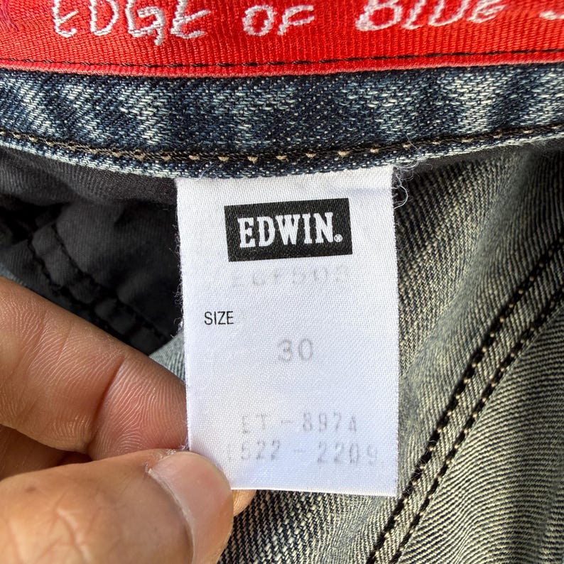 以下が含まれることがあります： デニム生地のクローズアップ。白いタグにはブランド名「Edwin」とサイズ「30」が表示されています。衣類のエッジには、赤い背景に白い文字で「EDGE OF BLUE」と書かれています。