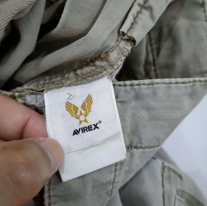 May include: Close-up of a tag with the brand name AVIREX&reg; and a gold emblem featuring wings and a star. The tag is attached to a pair of khaki-colored pants, with visible stitching and fabric texture.