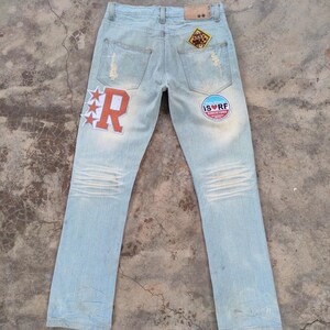 May include: Light blue denim jeans featuring a large orange letter R patch with three stars on the left leg, a circular patch with the text IS RF SINCE 1984, a diamond-shaped patch on the right pocket, and distressed detailing on the back pockets.