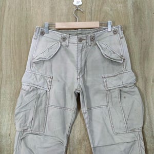 May include: Light tan cargo pants with multiple pockets and button closures. The pants are hanging on a wooden hanger. The stitching is a darker tan color.