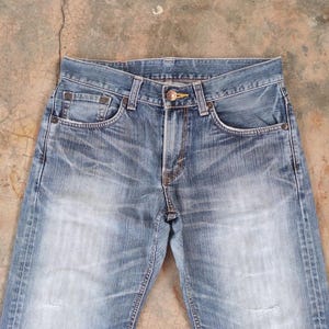 May include: Light blue denim jeans with a faded wash and distressed details. The jeans feature classic five-pocket styling, a button and zipper closure, and belt loops. The denim has a worn appearance.