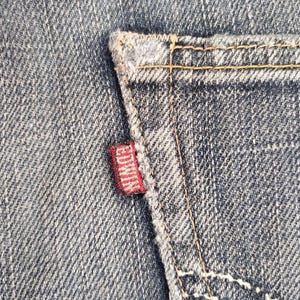 Puede incluir: Primer plano de una secci&oacute;n de vaqueros azules. Una peque&ntilde;a etiqueta roja con la marca "EDWIN" est&aacute; cosida en el bolsillo trasero. Las costuras son de color amarillo.