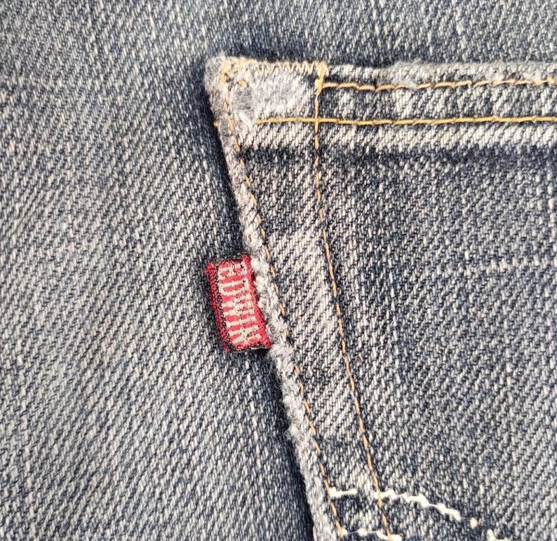 Puede incluir: Primer plano de una secci&oacute;n de vaqueros azules. Una peque&ntilde;a etiqueta roja con la marca "EDWIN" est&aacute; cosida en el bolsillo trasero. Las costuras son de color amarillo.