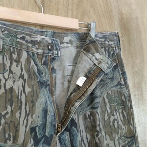 May include: Camouflage trousers with a button closure and a zip. The trousers are in shades of green, brown, and grey, with a pattern resembling tree bark. The trousers are hanging on a wooden hanger.