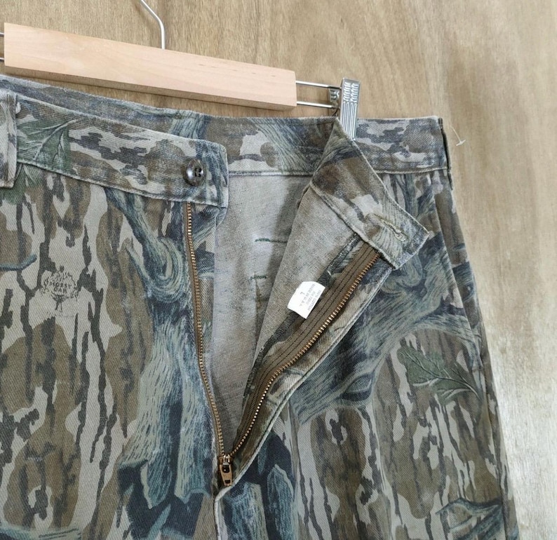 May include: Camouflage trousers with a button closure and a zip. The trousers are in shades of green, brown, and grey, with a pattern resembling tree bark. The trousers are hanging on a wooden hanger.