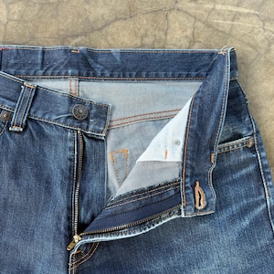 May include: Close-up of blue denim jeans with an open zipper and button closure. The jeans have a classic five-pocket design with copper-colored stitching. The fly is unbuttoned, revealing the light blue interior fabric.