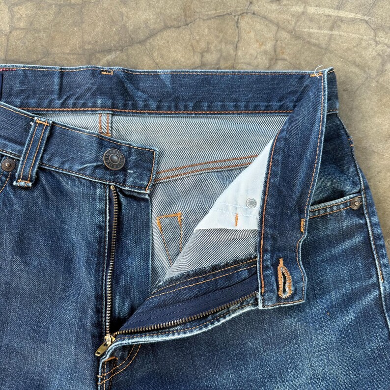 May include: Close-up of blue denim jeans with an open zipper and button closure. The jeans have a classic five-pocket design with copper-colored stitching. The fly is unbuttoned, revealing the light blue interior fabric.