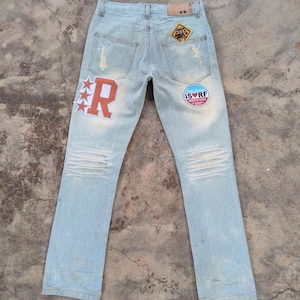 Könnte beinhalten: Hellblaue Jeans aus Denim mit Used-Details und Aufnähern. Zu den Merkmalen gehören ein "R"-Patch mit Sternen, ein kreisförmiger "ISURF"-Patch und ein kleiner quadratischer Patch in der Nähe der Gesäßtasche. Die Jeans haben ein klassisches Fünf-Taschen-Design.