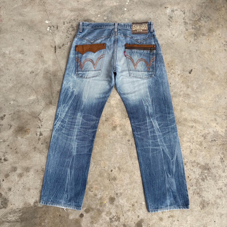 May include: Blue denim jeans with brown leather pocket accents and red stitching. The jeans have a faded wash and a leather patch on the back. The back pockets feature a distinctive red stitch design.