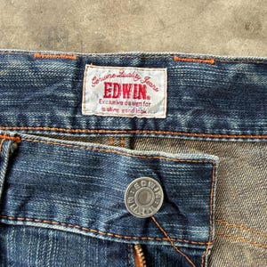 Pu&ograve; includere: Primo piano di jeans blu in denim con bottone e cerniera visibili. I jeans presentano un'etichetta del marchio con la scritta "EDWIN" e "Genuine Quality Jeans". Le cuciture sono di colore arancione a contrasto.