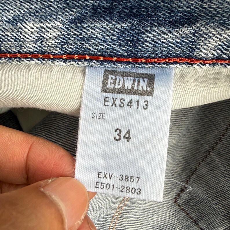 May include: Close-up of a denim garment tag with the brand name "EDWIN" in a rectangular box. The tag displays "EXS413" and the size "34". Additional codes "EXV-3857" and "E501-2803" are also visible.