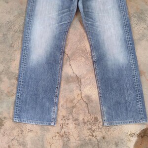 May include: Faded blue denim jeans with a straight leg cut. The jeans have a classic five-pocket design and a button and zipper closure. The denim has a worn look with lighter areas on the thighs and knees.