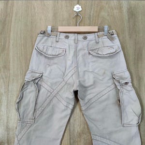 May include: Khaki-colored cargo pants with multiple pockets and decorative stitching. The pants are hanging on a wooden hanger against a light brown wooden background. The pants have a button and zipper closure.