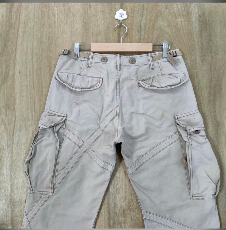 May include: Khaki-colored cargo pants with multiple pockets and decorative stitching. The pants are hanging on a wooden hanger against a light brown wooden background. The pants have a button and zipper closure.