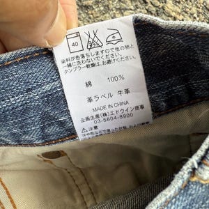 May include: Close-up of a denim garment tag with Japanese text and care symbols. The tag indicates the fabric is 100% cotton and features a leather label. Washing instructions are also included.