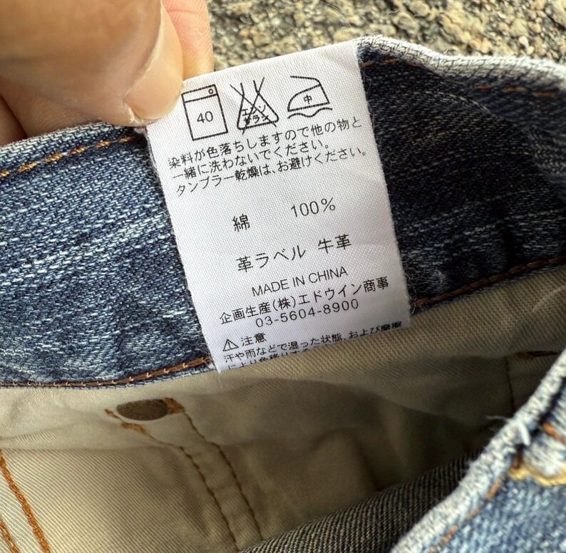May include: Close-up of a denim garment tag with Japanese text and care symbols. The tag indicates the fabric is 100% cotton and features a leather label. Washing instructions are also included.