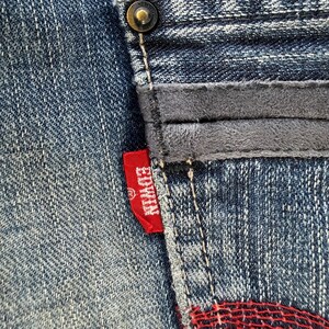 以下が含まれることがあります： デニム生地のクローズアップ。グレーのスエードのようなパッチと赤いEdwinタグが付いています。ジーンズはボタン留めで、ステッチが見えます。デニムは色あせた青色です。