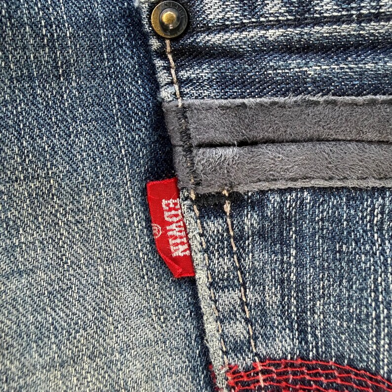 以下が含まれることがあります： デニム生地のクローズアップ。グレーのスエードのようなパッチと赤いEdwinタグが付いています。ジーンズはボタン留めで、ステッチが見えます。デニムは色あせた青色です。