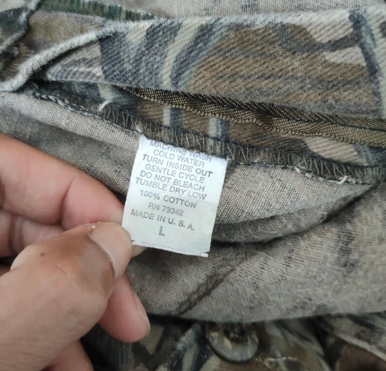 May include: Close-up of a camouflage-patterned fabric with a care label. The label reads "MACHINE WASH COLD WATER TURN INSIDE OUT GENTLE CYCLE DO NOT BLEACH TUMBLE DRY LOW 100% COTTON RN 73342 MADE IN U. S. A. L".