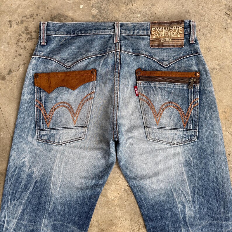 May include: Light blue denim jeans with brown leather pocket accents and orange stitching. The back pockets feature a unique wave design. The jeans have a faded wash and a leather patch that reads "EXCLUSIVE VINTAGE EDWIN".