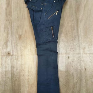 Pu&ograve; includere: Pantaloni cargo blu navy scuro con pi&ugrave; tasche e cerniere dorate. I pantaloni sono appesi a un'appendiabiti su uno sfondo di legno chiaro. I pantaloni hanno un design a gamba dritta.