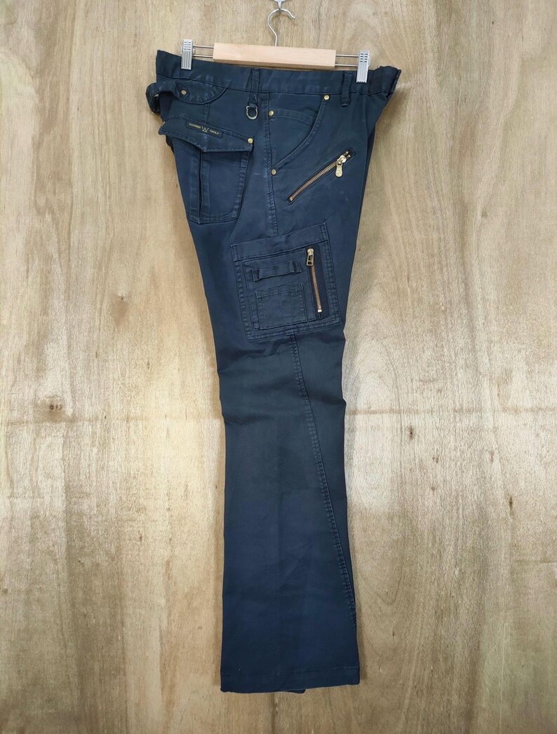 Pu&ograve; includere: Pantaloni cargo blu navy scuro con pi&ugrave; tasche e cerniere dorate. I pantaloni sono appesi a un'appendiabiti su uno sfondo di legno chiaro. I pantaloni hanno un design a gamba dritta.