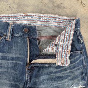 May include: Close-up of blue denim jeans with a partially unzipped fly. The jeans feature a decorative patterned lining in shades of blue, red, and white. The button is visible, and the jeans appear worn.