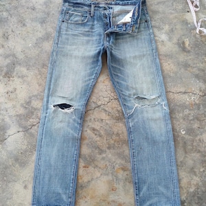 Könnte beinhalten: Hellblaue Jeans mit Used-Details, einschließlich zerrissener Knie. Die Jeans haben ein klassisches Fünf-Taschen-Design und einen Knopf- und Reißverschluss. Die Jeans liegen flach auf einer Betonoberfläche.