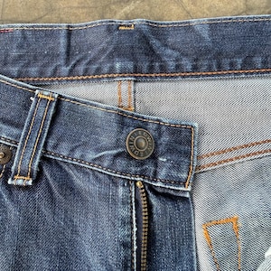 May include: Close-up of a pair of blue denim jeans. The image highlights the details of the jeans, including the zipper, button, and stitching. The button has the brand name "EDWIN" engraved on it.